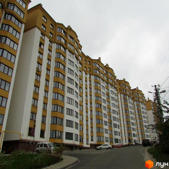 Житловий комплекс Щасливий на Петропавлівці. Фасад будинку біло-жовтого кольору з великою кількістю вікон. Поруч дорога та зелені насадження.