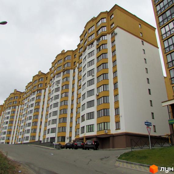 Житловий комплекс Щасливий на Петропавлівці - багатоповерхова новобудова з жовто-білим фасадом та численними вікнами. Поруч розташована прибудинкова територія з паркуванням автомобілів.