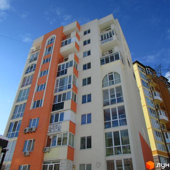 Житловий комплекс Квітневий: фасад будинку з помаранчевими та білими елементами, великою кількістю вікон та балконів. Вид на будинок знизу на фоні блакитного неба.