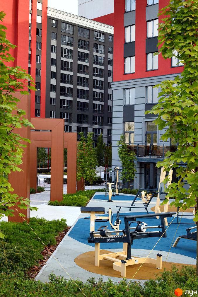 Житловий комплекс Irpin City з червоними та сірими фасадами. На прибудинковій території розташований сучасний спортивний майданчик з тренажерами.
