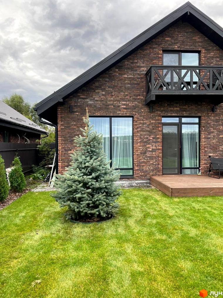 Цегляний будинок з балконом та великими вікнами, перед будинком газон та блакитна ялинка. Містечко котеджів Бельгійське село.