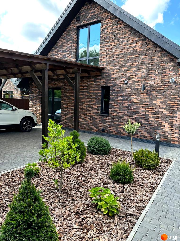 Цегляний будинок з темною покрівлею та великими вікнами. На прибудинковій території висаджені дерева та кущі, а також викладена бруківка.