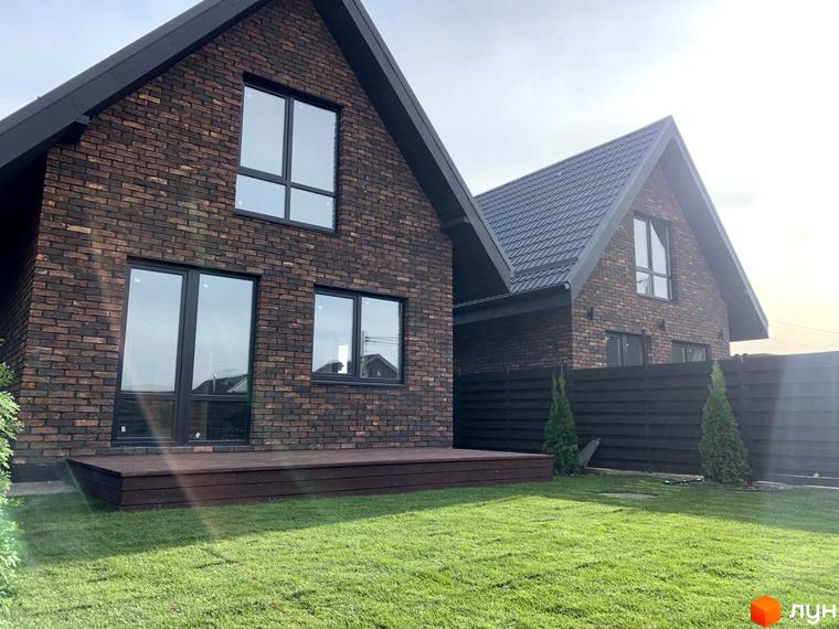 Цегляні будинки з панорамними вікнами та темною покрівлею в котеджному містечку. На території газон та декоративні дерева, огородження.