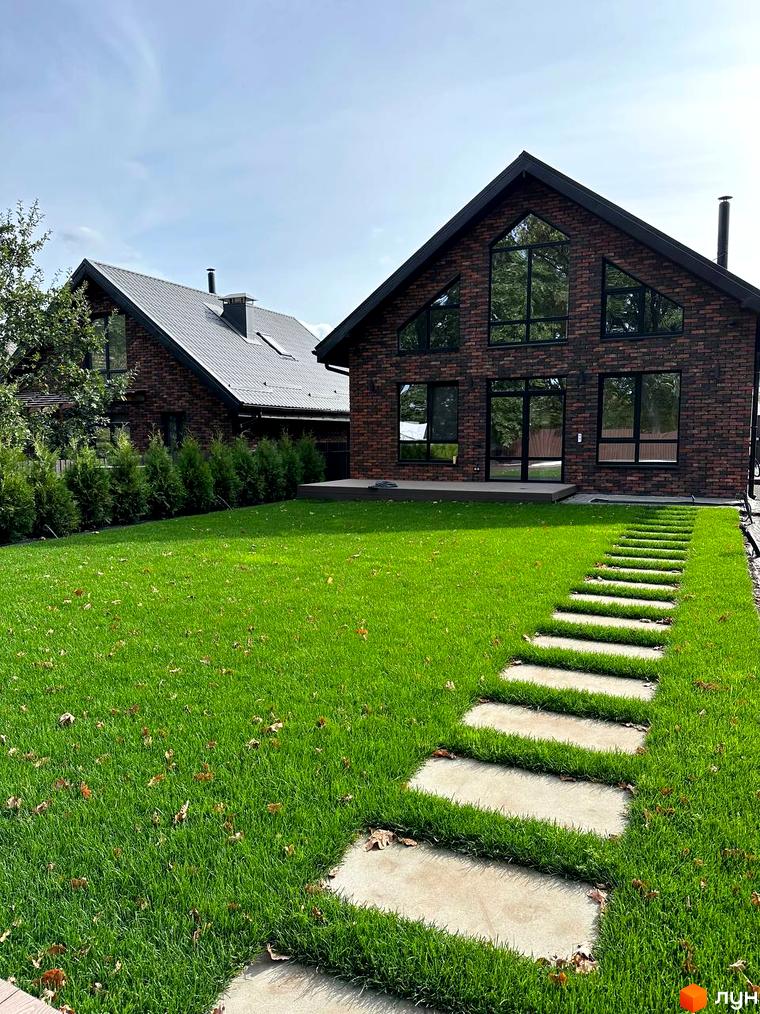 Цегляний будинок з великими вікнами та чорним дахом. Доглянутий газон з кам'яною доріжкою веде до будинку. КМ Бельгійське село.