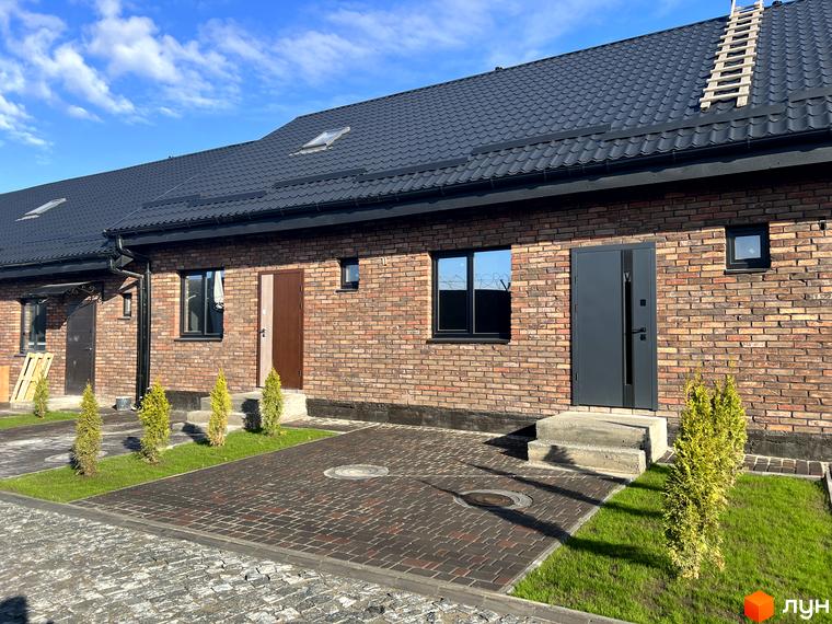 Цегляний будинок з темною покрівлею та вхідними дверима. Перед будинком бруківка, газон та декоративні рослини, що створюють затишну атмосферу.