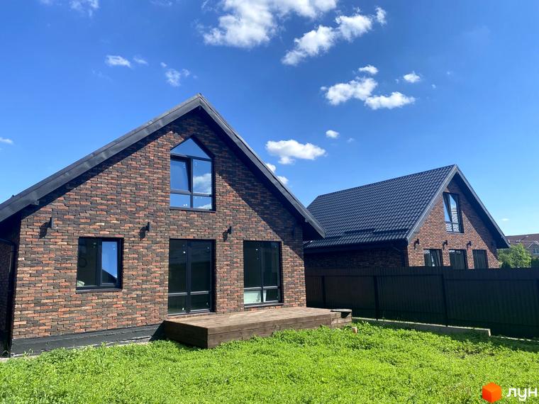 Цегляний будинок з великими вікнами та дерев'яною терасою в Містечко котеджів Бельгійське село. Навколо будинку зелена трава та блакитне небо з хмарами.