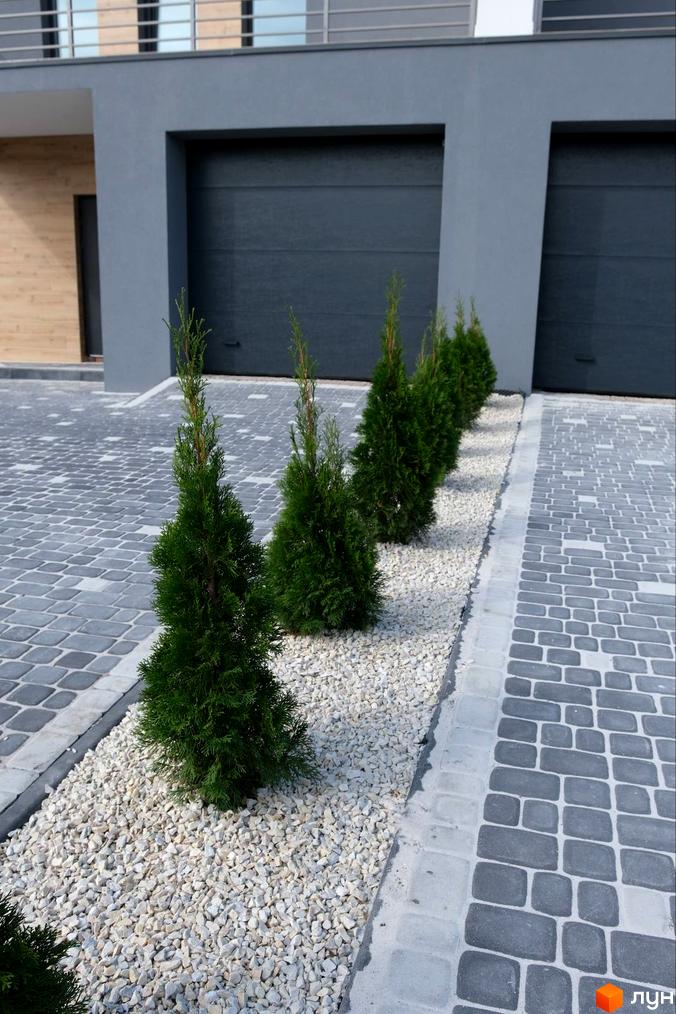 Сірий будинок з темними гаражними воротами та доріжка з бруківки з рядом туй на білому гравії.