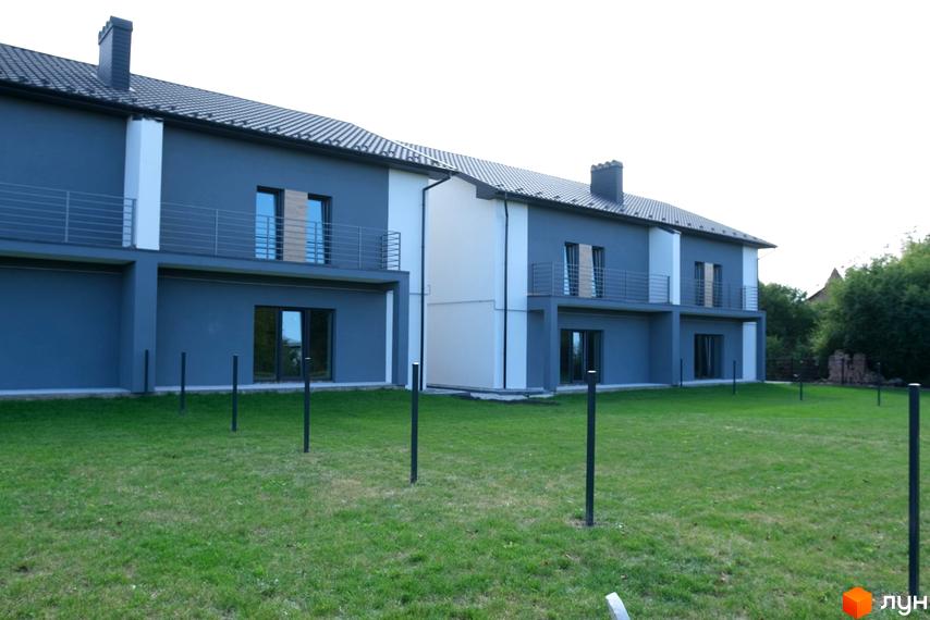 Зображення житлового комплексу: двоповерховий будинок з сірим фасадом, білими вставками, балконами та зеленим газоном.