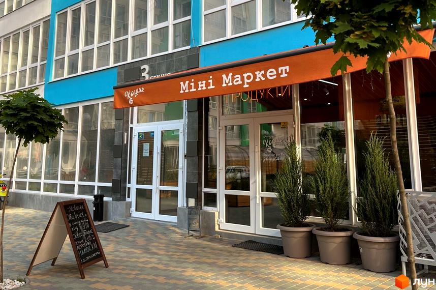 Фасад будівлі житлового комплексу з магазином на першому поверсі. Біля входу в магазин стоять вазони з рослинами та рекламна вивіска.