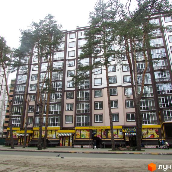 Зовнішній вигляд новобудови ЖК Парковий з великою кількістю вікон та балконів, оточений високими соснами. На першому поверсі розташовані магазини та комерційні приміщення.