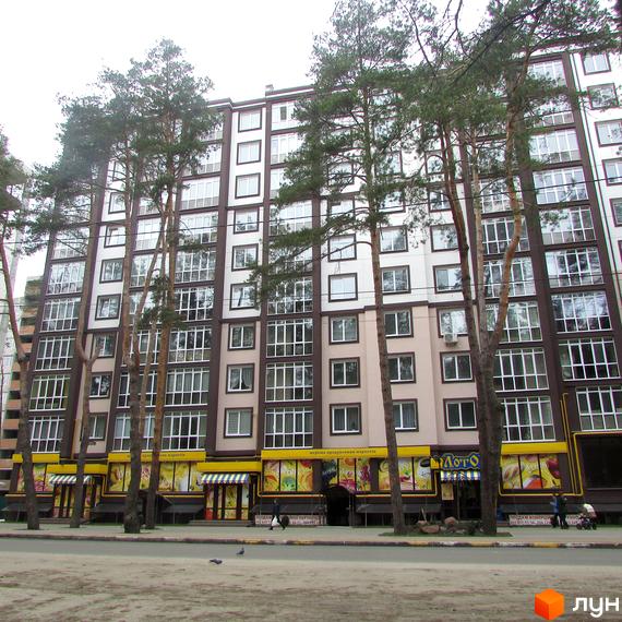Зовнішній вигляд новобудови ЖК Парковий з великою кількістю вікон та балконів, оточеної високими деревами. На першому поверсі розташовані комерційні приміщення з яскравими вивісками.