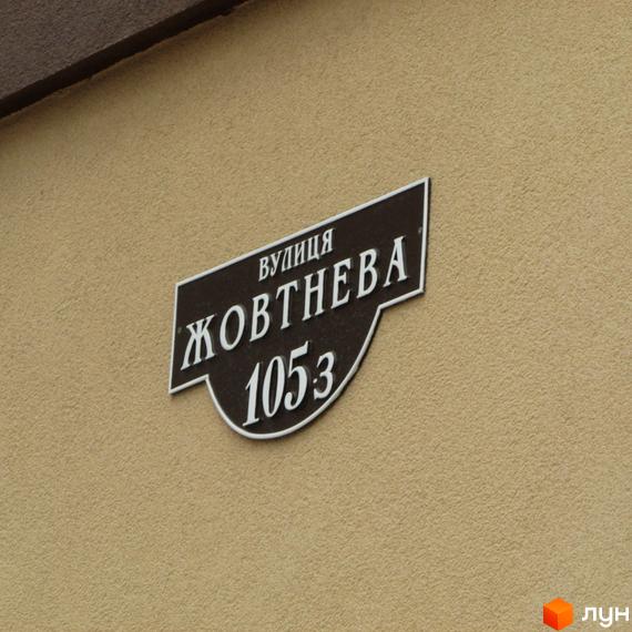 Вуличний знак з назвою вулиці Жовтнева та номером будинку 1053 на фасаді будівлі. Фасад будівлі виконаний у світлих тонах з текстурованою поверхнею.