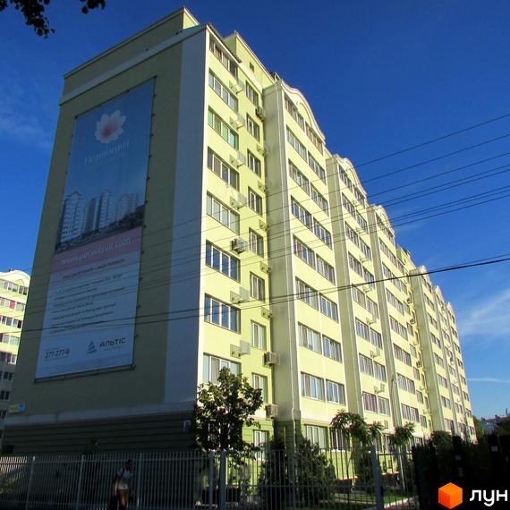 Висотна будівля ЖК Первоцвіт з великою рекламною вивіскою на фасаді. На прибудинковій території видно огорожу та зелені насадження.