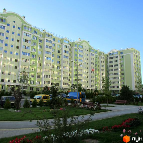 Висотна будівля Житлового комплексу Первоцвіт з великою кількістю вікон та світлим фасадом. На прибудинковій території є зелені насадження, парковка та доріжки для прогулянок.