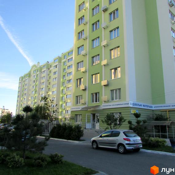 Зображення ЖК Первоцвіт. Багатоповерховий будинок з зеленими стінами та білими вставками. На передньому плані дорога з припаркованим автомобілем та зелені насадження.