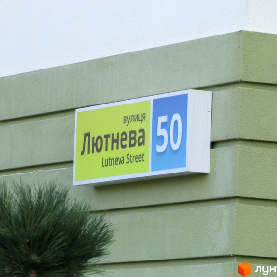Вивіска з назвою вулиці Lutneva Street та номером будинку 50 на фасаді будівлі. Зелений колір стін та зелень на передньому плані.