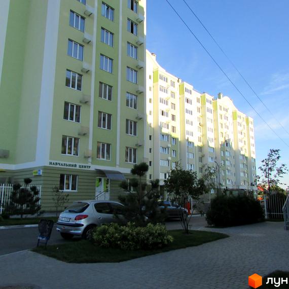 Зображення житлового комплексу Первоцвіт. Багатоповерховий будинок з великою кількістю вікон, світлого кольору. На прибудинковій території є зелені насадження та парковка.