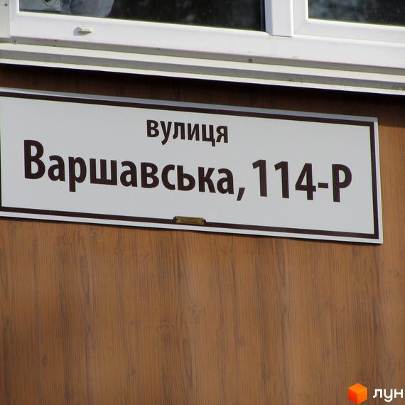 Вуличний знак з написом «Варшавська, 114-Р». Напис розташований на білій табличці з коричневою рамкою, що кріпиться на стіні з дерев'яним покриттям.