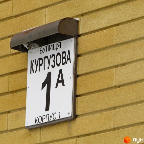 Вивіска з назвою вулиці Кургузова, номер будинку 1А, корпус 1. Це частина фасаду будинку з жовтою цегляною кладкою.