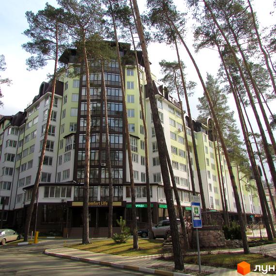 Житловий комплекс Ірпінські Липки - багатоповерхова новобудова світло-зеленого та білого кольору з великою кількістю вікон, оточена високими соснами. На прибудинковій території видно дорогу, тротуар та зелені насадження.