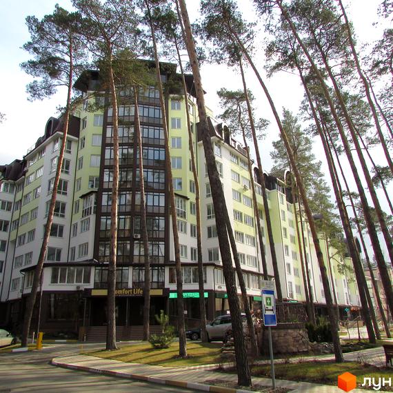 Житловий комплекс Ірпінські Липки - багатоповерховий будинок світло-зеленого та білого кольорів з великою кількістю вікон, оточений соснами. Поруч розташована доглянута прибудинкова територія з тротуаром та газоном.