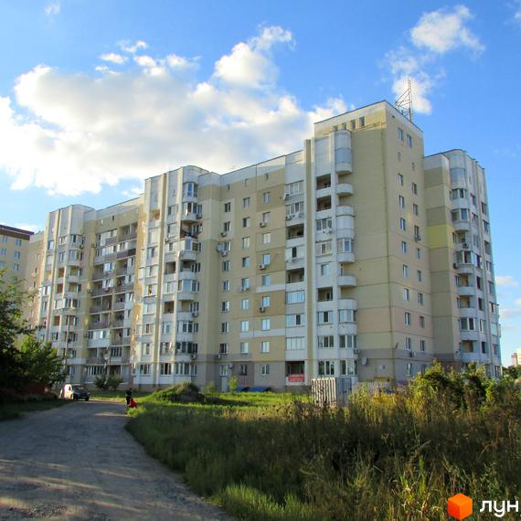 Багатоповерховий житловий комплекс Уюткіно з балконами та кондиціонерами на фоні блакитного неба з хмарами. Прибудинкова територія з зеленою травою та дорогою.