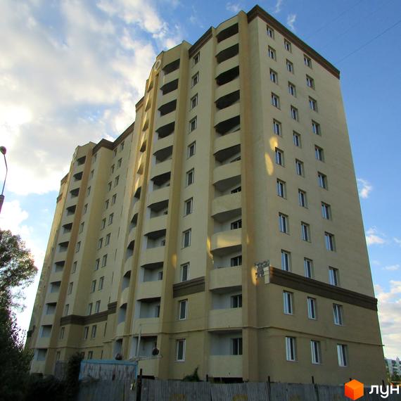 Житловий комплекс Уюткіно - висотна новобудова з балконами та вікнами на тлі блакитного неба з хмарами. Навколо будинку видно паркан та дерева.