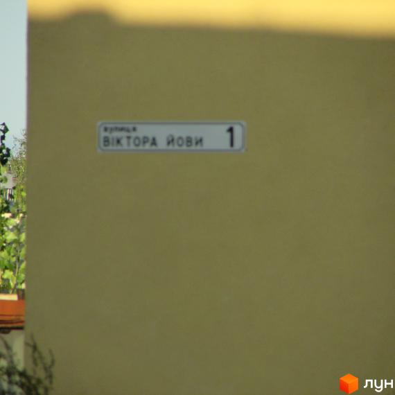 На фасаді будинку табличка з назвою вулиці Віктора Йови та номером 1. Навколо зелені дерева та рослини.