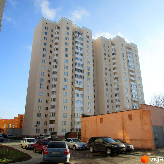 Високий світло-бежевий житловий комплекс з численними балконами та кондиціонерами. На прибудинковій території розташовані припарковані автомобілі.