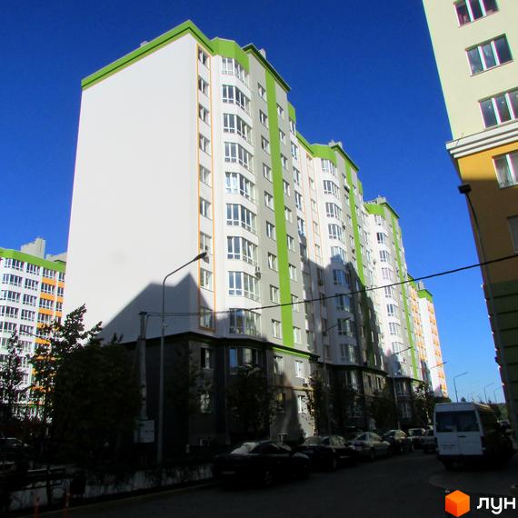 Житловий комплекс Евромісто - багатоповерхові будинки з білими та зеленими фасадами, заскленими балконами, на тлі блакитного неба. Біля будинку дорога з припаркованими автомобілями та деревами.
