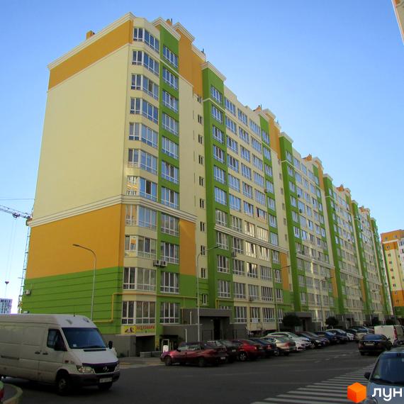 Зовнішній вигляд багатоповерхового житлового комплексу Евромісто з яскравим фасадом, що складається з жовтих, світло-бежевих та зелених секцій. Перед комплексом розташована парковка з автомобілями та пішохідний перехід.