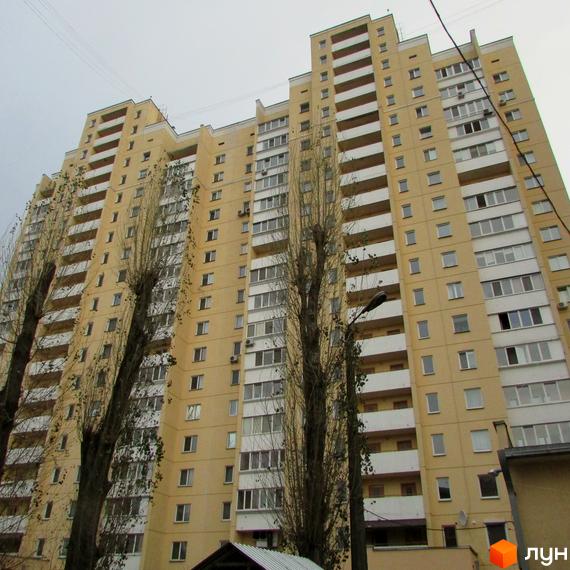Високий житловий будинок бежевого кольору з численними білими балконами та вікнами. На передньому плані видно голі дерева та вуличний ліхтар.