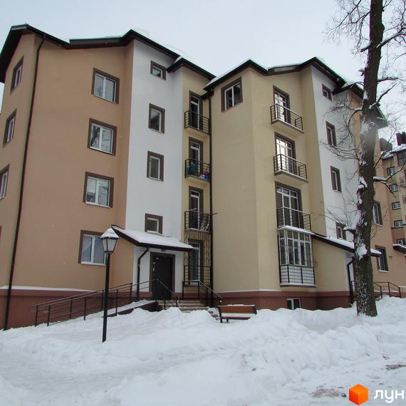 Зовнішній вигляд житлового комплексу Джерело взимку. Будинок складається з кількох секцій різної висоти, оздоблених світлою та бежевою штукатуркою. Перед будинком розташована засніжена прибудинкова територія з лавкою.