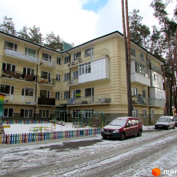 Житловий комплекс на вул. Рильського, 7, світло-жовтого кольору з балконами та зеленим дахом. Перед будинком засніжений дитячий майданчик та припарковані автомобілі.