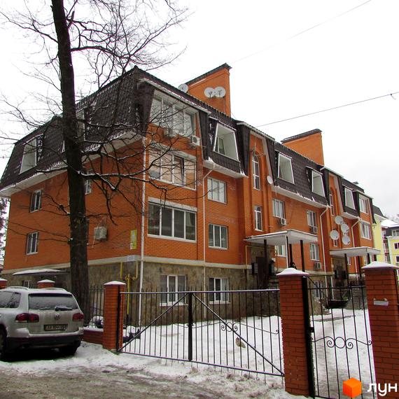 Зовнішній вигляд цегляного будинку з мансардними вікнами та супутниковими антенами. Прибудинкова територія огороджена парканом, на подвір'ї видно автомобіль та сніг.