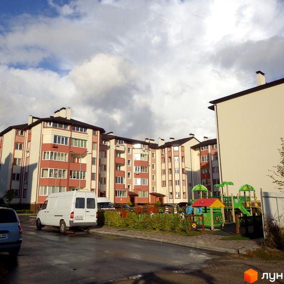 Житловий комплекс Щасливий з червоно-білими фасадами та дитячим майданчиком. На території комплексу є парковка та зелені насадження.