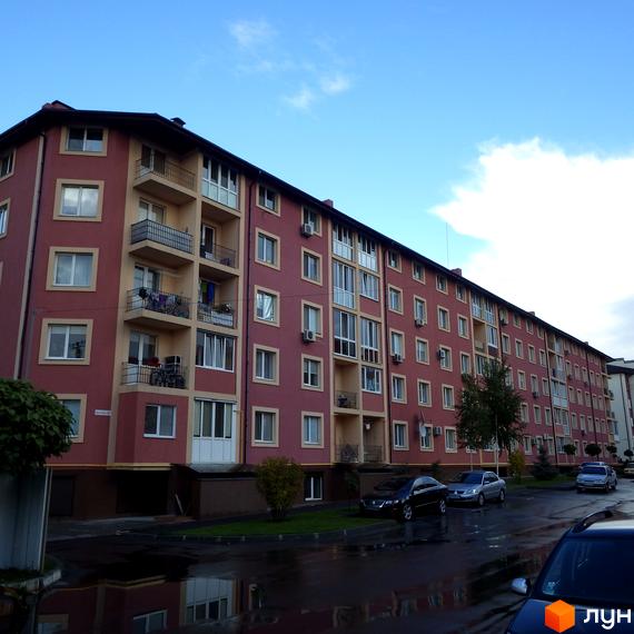 Зовнішній вигляд багатоповерхового будинку ЖК Щасливий з червоним фасадом та балконами. На прибудинковій території розташовані автомобілі та зелені насадження.