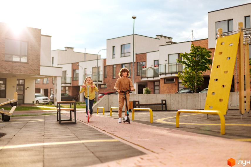 Котеджне містечко КМ Липові Роси: діти катаються на самокатах біля сучасних будинків з цегляним фасадом. Прибудинкова територія з дитячим майданчиком.