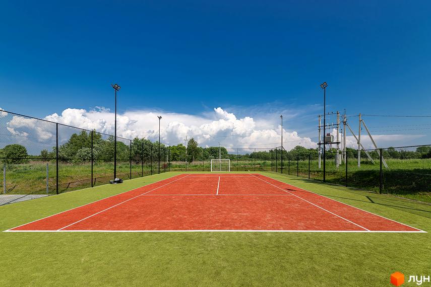 Котеджне містечко Липові Роси: спортивний майданчик з червоним покриттям та зеленою зоною навколо, оточений парканом, на фоні ясного неба з хмарами.