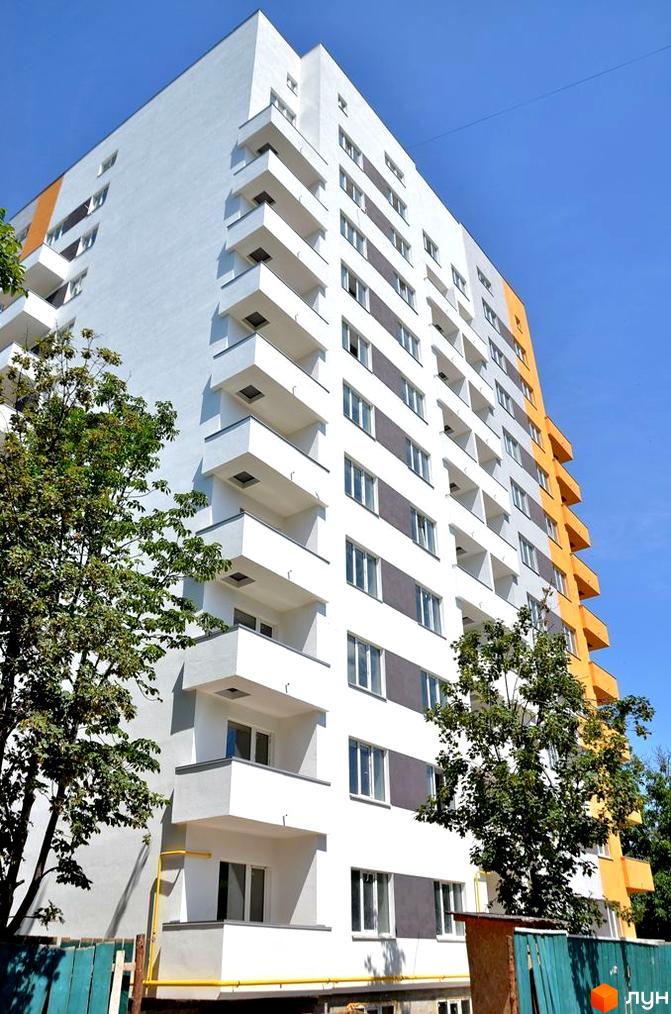 Зовнішній вигляд новобудови ЖК Щасливий у Вишневому. Будинок має біло-помаранчеву фасадну частину з численними балконами та вікнами. Навколо будинку зелені дерева та елементи прибудинкової території.