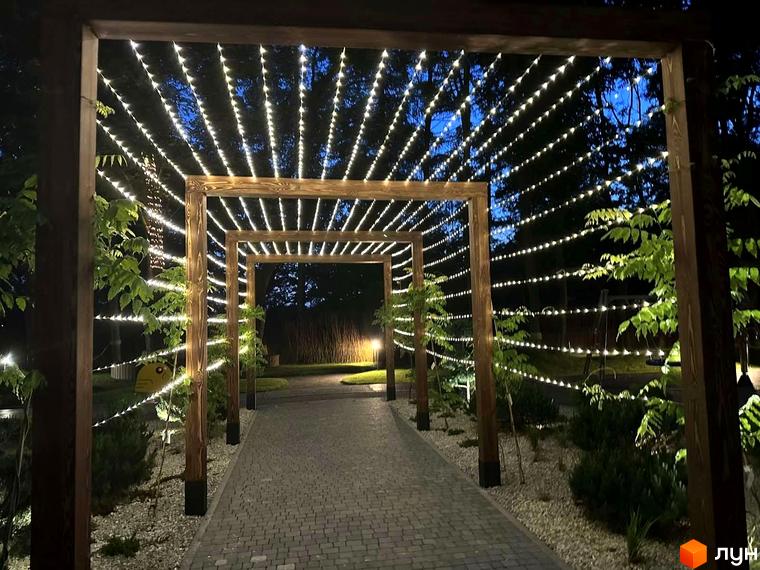 Вхідна арка з гірляндами в ЖК Tiffany веде до доглянутої території з бруківкою та зеленими насадженнями.