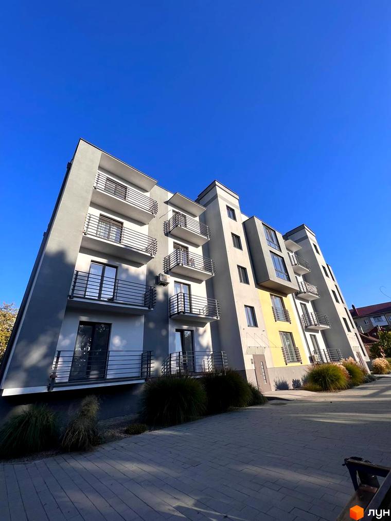 Висотна будівля ЖК Сучасний з балконами та панорамними вікнами на тлі блакитного неба. На передньому плані бруківка та зелені насадження.