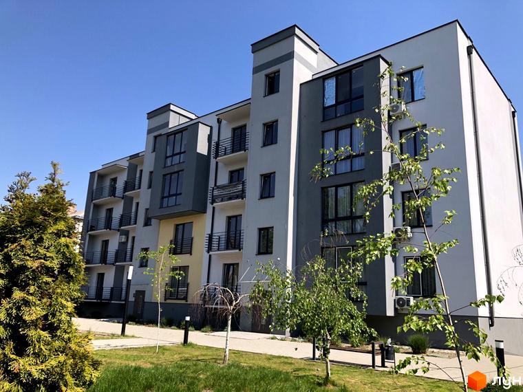 Багатоповерхова новобудова з панорамними вікнами та балконами. На прибудинковій території газон, дерева та доріжки для прогулянок.