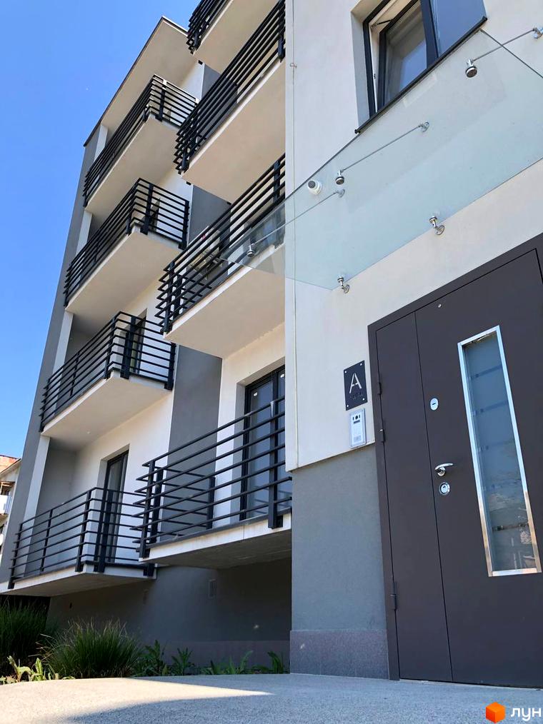 Багатоповерховий будинок ЖК Сучасний з балконами та металевими поручнями. Біля входу скляний навіс та двері з вікном, на території зелені насадження.