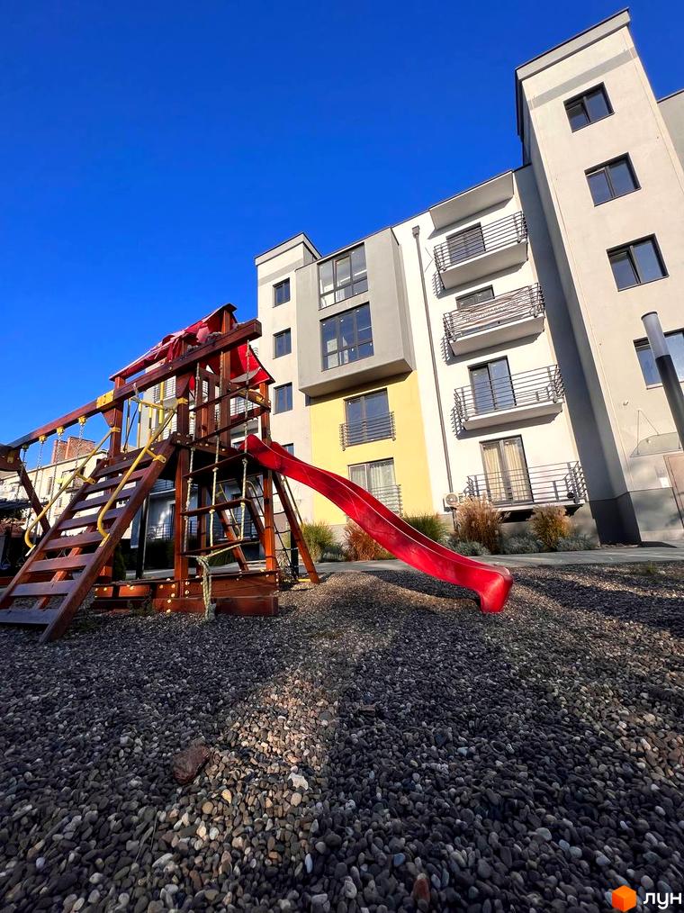 Новобудова з балконами та вікнами на тлі блакитного неба. Поруч дитячий майданчик з гіркою та сходами.