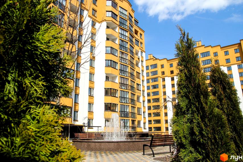 Житловий комплекс Щасливий на Петропавлівці. Фасад будинку жовто-білого кольору, з балконами, на прибудинковій території фонтан та лавки.