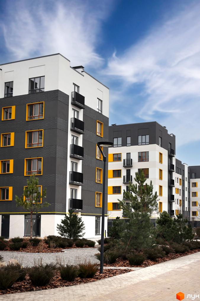 Житловий комплекс Молодість: фасади будівель з сірими та білими елементами, жовтими вікнами та балконами. Прибудинкова територія з деревами та кущами.