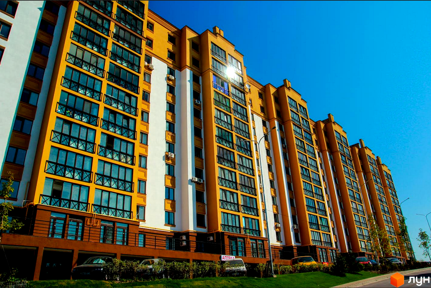 Житловий комплекс Щасливий на Петропавлівці – це багатоповерховий будинок жовто-білого кольору з численними вікнами та балконами. Навколо будинку розташована зелена прибудинкова територія з деревами та автомобілями.