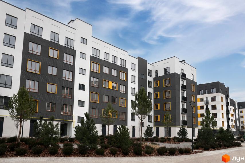 Фасад житлового комплексу Молодість з білими та темно-сірими елементами та жовтими акцентами на вікнах. Прибудинкова територія з молодими деревами та кущами.