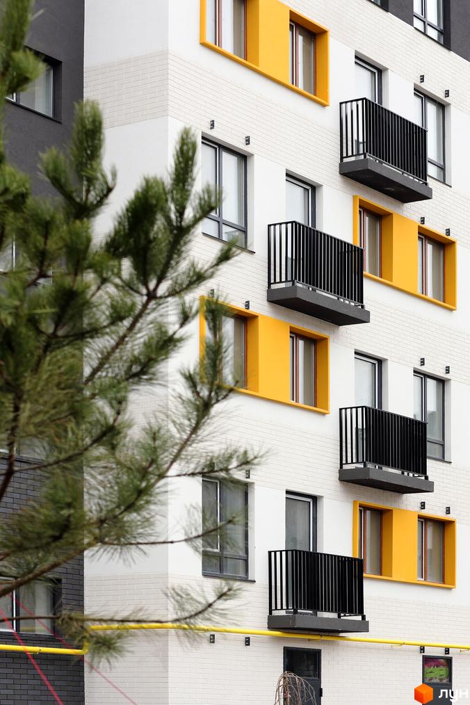 Фасад житлового комплексу Молодість з жовтими елементами та чорними балконами. На передньому плані видно гілки хвої.